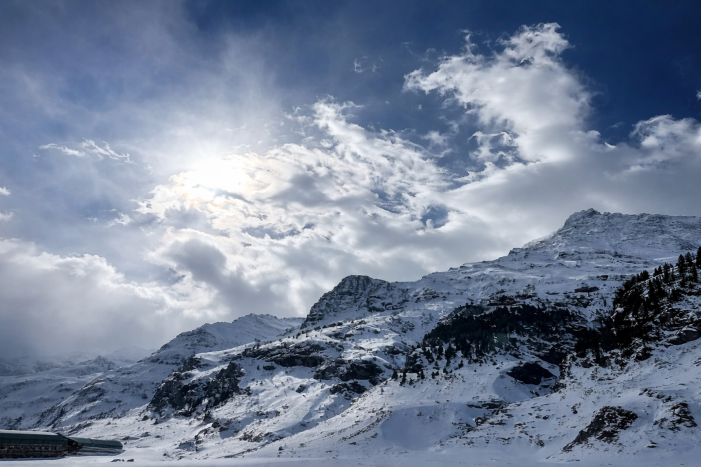 A wide scenic view of snow-capped Himalayan mountains under a bright sun and cloudy sky.